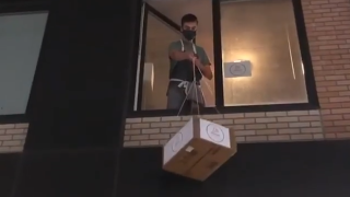 Masked man lowers cardboard box out of window using ropes.