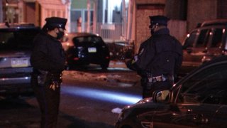 Two police officers, wearing hats and face masks, stand next to one another as the officer to the right shines a light on the ground. Parked cars can be seen around them as they investigate a stabbing in the Overbrook neighborhood.