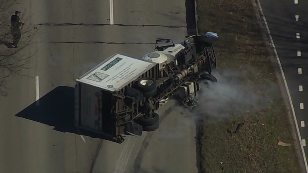 A box truck rests on its side as smoke wafts from it. The truck lays across multiple lanes of traffic.