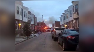 Cars are parked on a street line by row homes as a smoke and flames rise from a fire over the horizon.