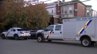 A Philadelphia police SUV and a small police truck park on a street in Philadelphia. To the side of the vehicles can be seen a tree in front of a pair of row homes.