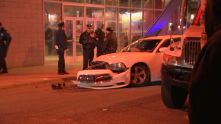 Philadelphia police officers stand on the sidewalk behind a white Dodge Charger with front-end bumper damage.