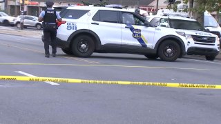 A police officer stands next to a police SUV parked on a street behind crime scene tape.