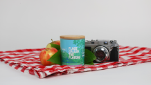 Place Called the Plateau candle with apples and a camera on a picnic blanket.