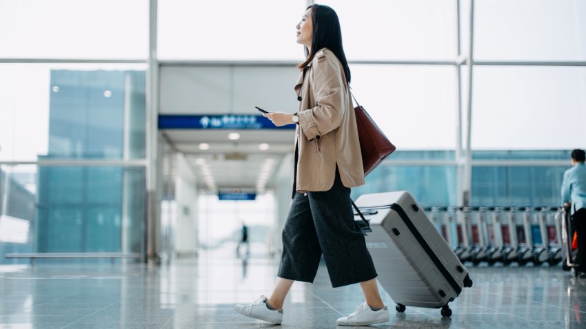 Young Asian woman carrying suitcase and holding smartphone on hand, walking in airport terminal. Ready to travel. Travel and vacation concept. Business person on business trip