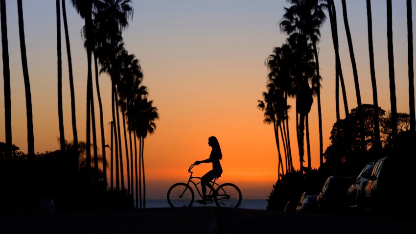 A woman rides a beach cruiser bicycle along a palm tree-lined street near the beach on a warm, fall day in San Diego’s La Jolla neighborhood at sunset.